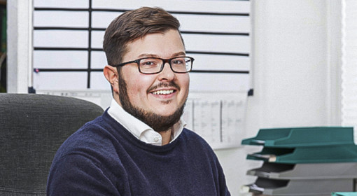 Junger Mann mit Brille und Bart sitzt im Büro und lächelt in die Kamera